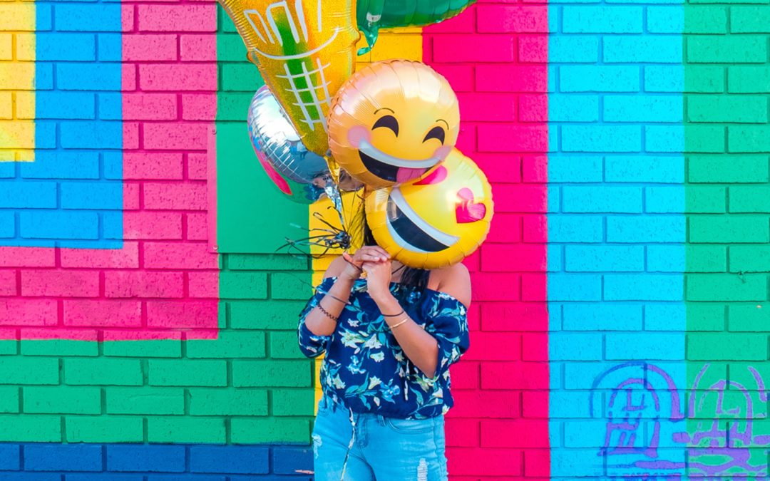 Person holding emoji balloons obscuring face standing in front of brightly painted exterior wall