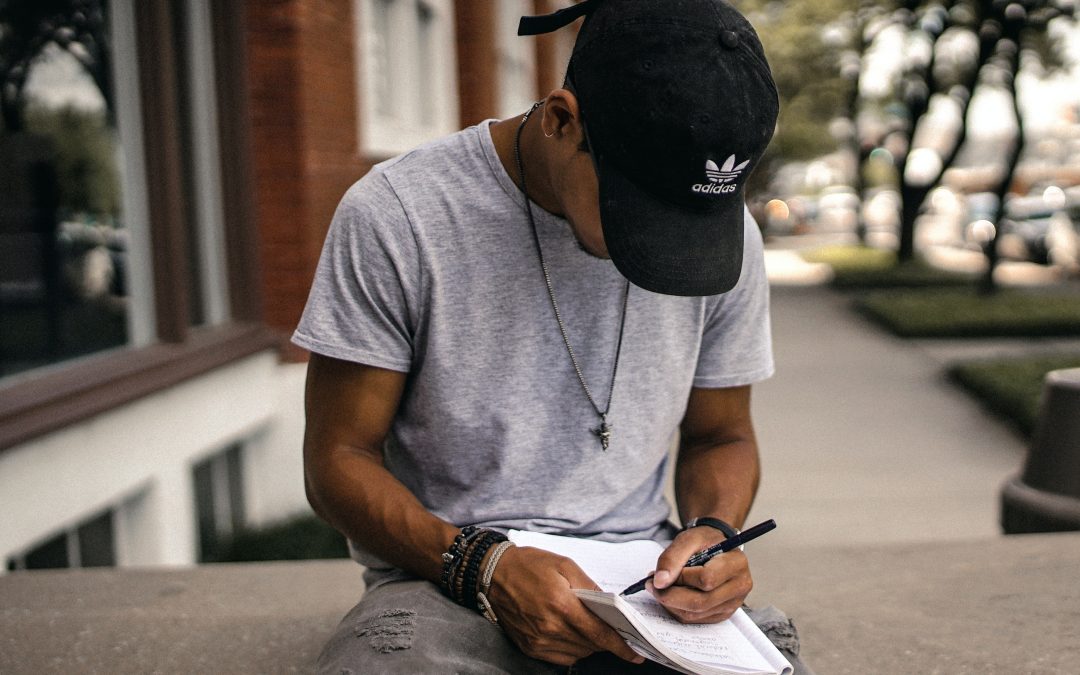 Man in a baseball cap and t-shirt sitting outside a shop on a bench engrossed in writing something on a pad of paper