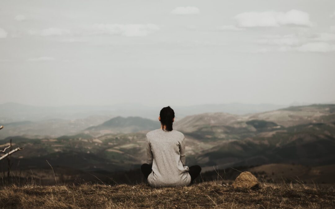 A woman sitting on a hill looking out at the landscape and thinking.
